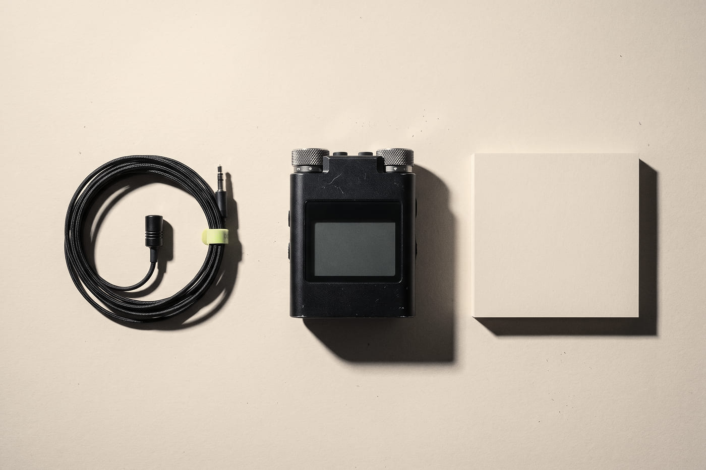 A field audio recorder, a coiled lavalier microphone, and a squared stack of blank forms